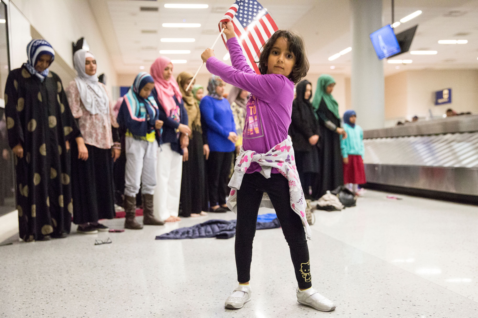 A young girl dances with an American flag in baggage claim while women pray behind her during a protest at the Dallas/Fort Worth International Airport. A young girl dances with an American flag in baggage claim while women pray behind her during a protest at the Dallas/Fort Worth International Airport.