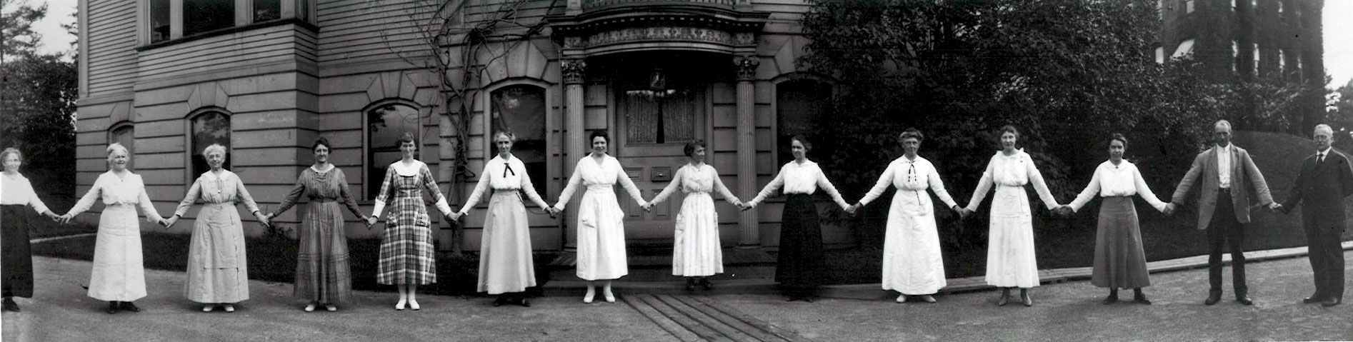 A dozen women hold hands in front of a building on the Harvard campus in 1918. They are all women "computers" working in the Harvard College Observatory. At the far right of the image is Edward Pickering, the head of the Observatory who hired them all.