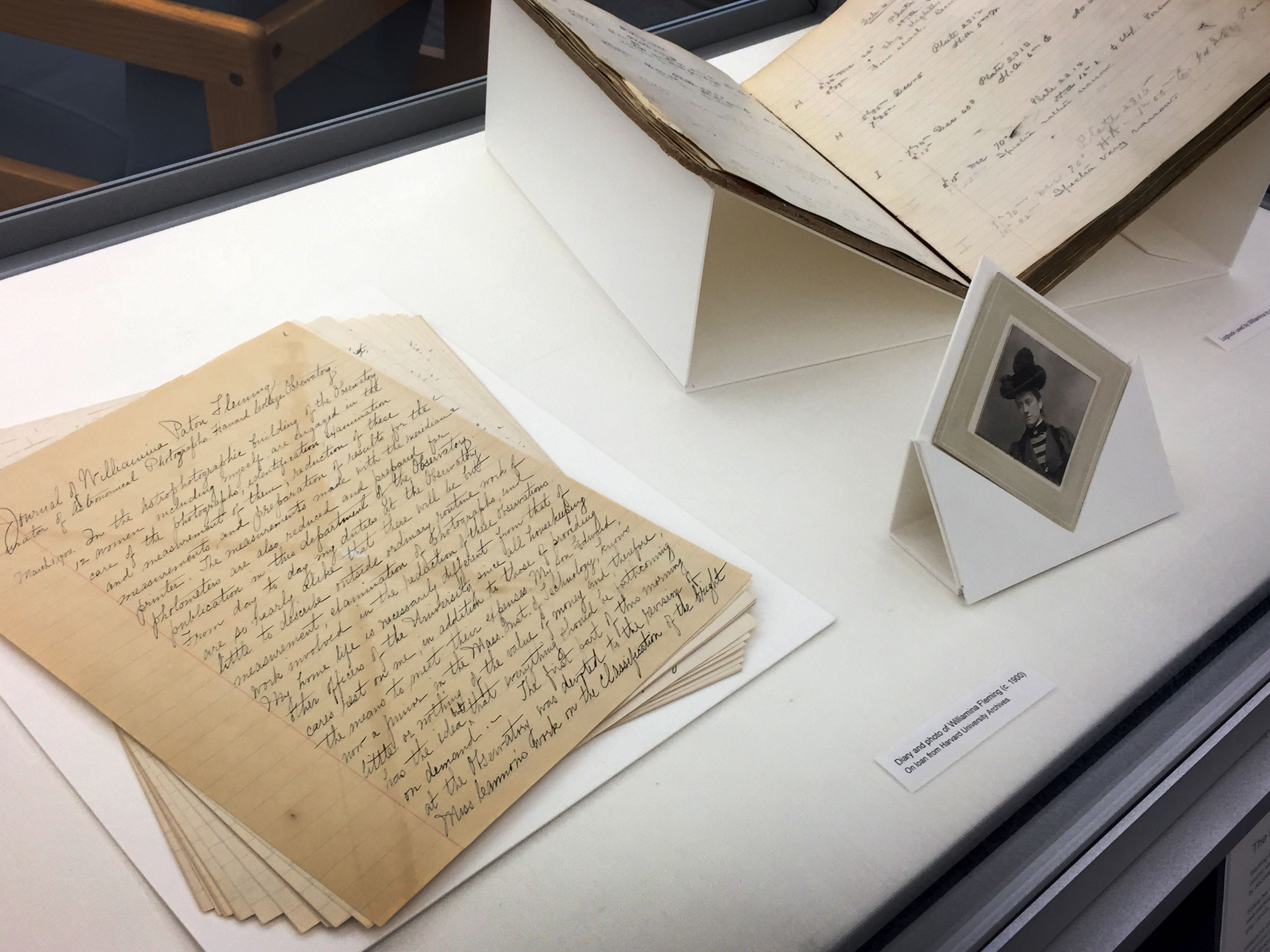 From left to right in a glass display case: yellowed pages with elegant handwritten script, a black and white portrait of Wilhelmina Fleming wearing a feathered hat; a yellowed logbook with mathematical notations in it. 