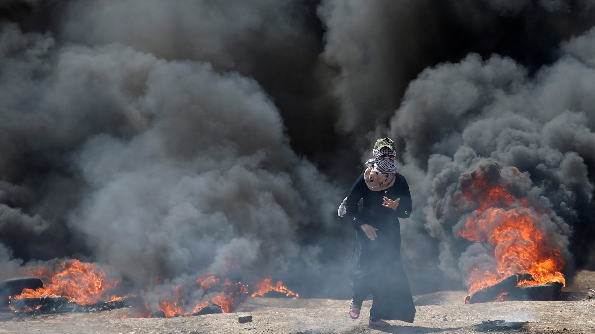 A Palestinian demonstrator gestures during a protest against US embassy move to Jerusalem at the Israel-Gaza border, May 14, 2018. A Palestinian demonstrator walks with black smoke and tires burning in the background.