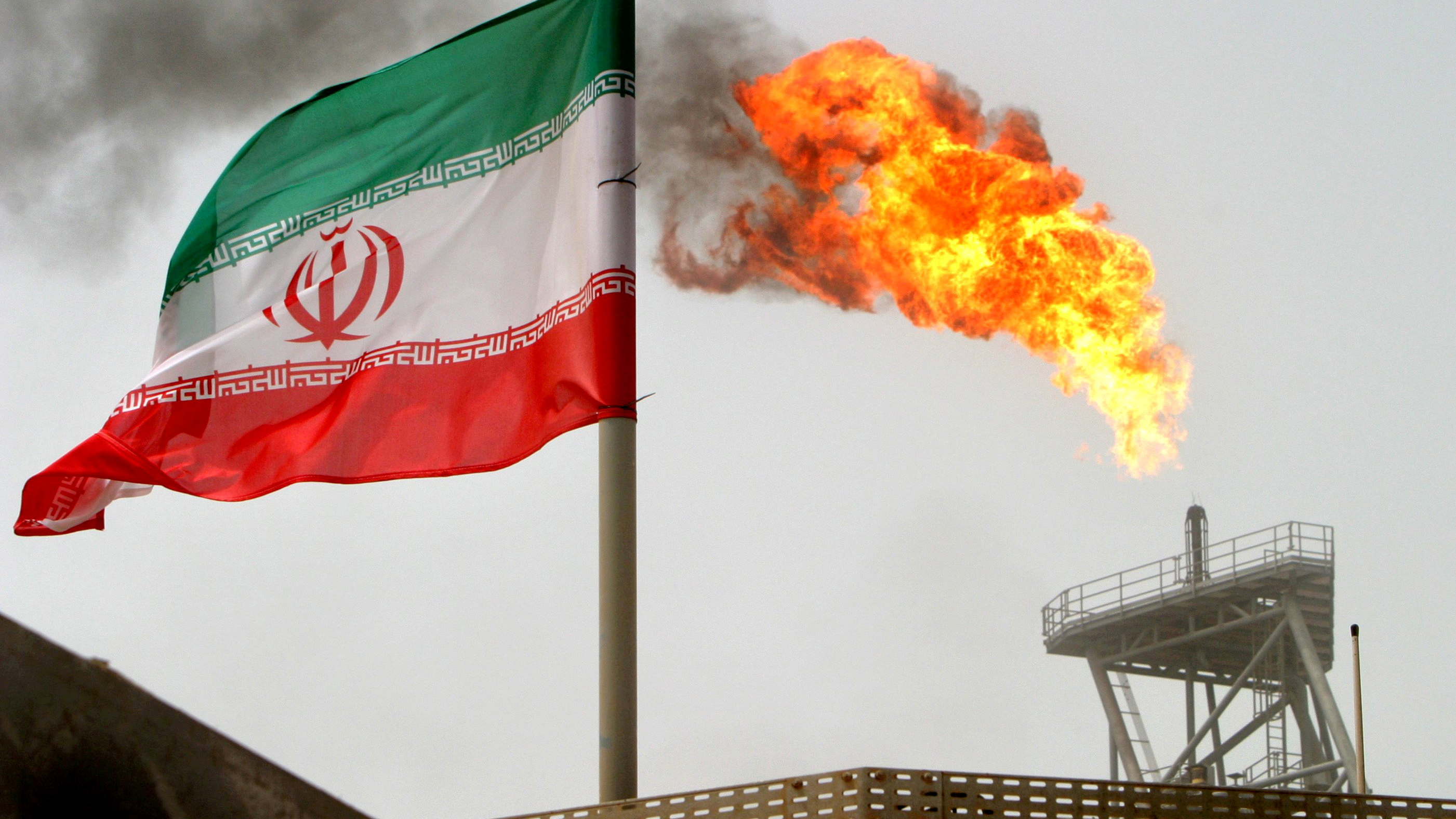 A gas flare on an oil production platform in the Soroush oil fields is seen alongside an Iranian flag in the Persian Gulf, Iran. A gas flare is seen in the background with the Iranian flag flying in the wind in the foreground.