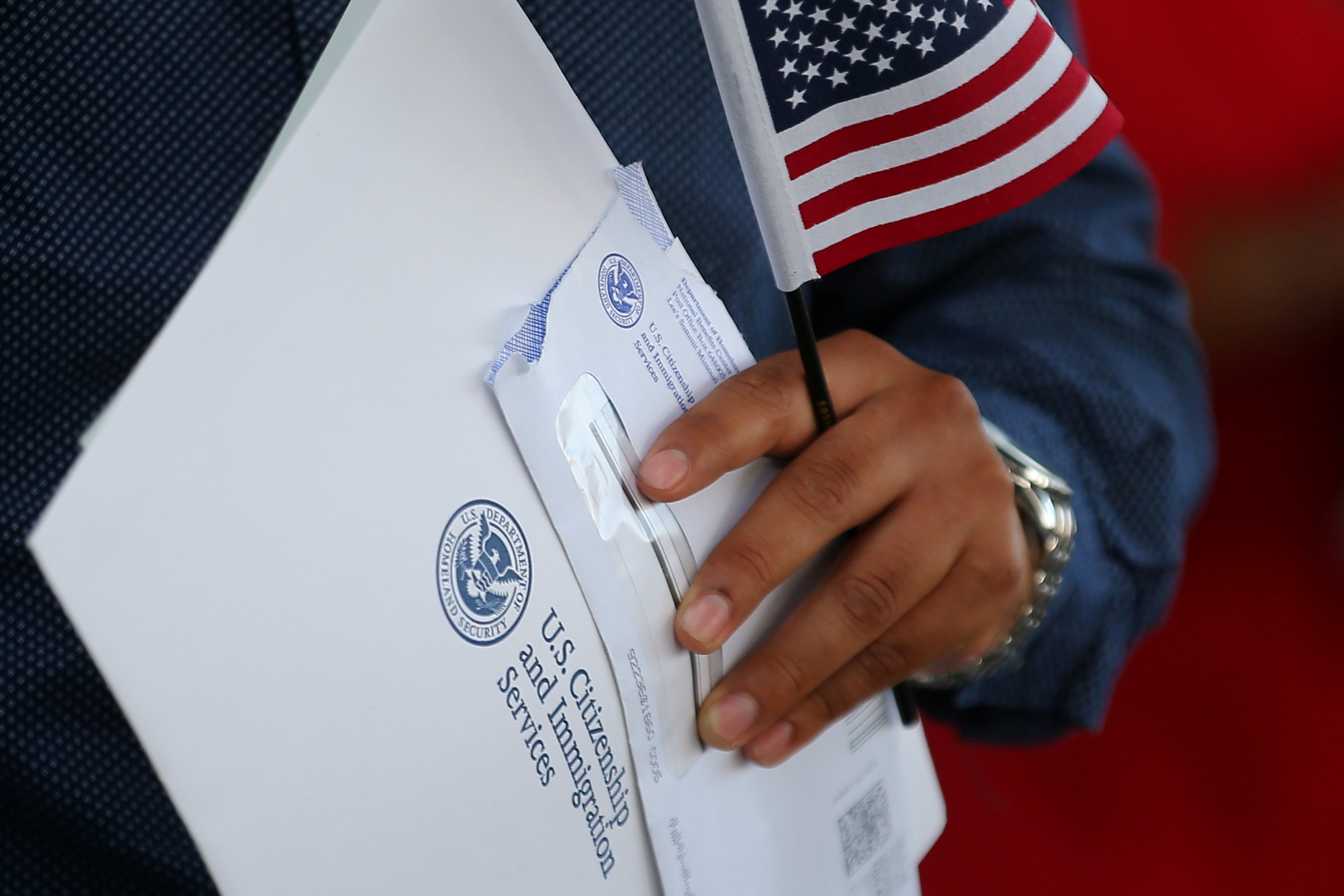 Close-up photo of man's hand, holding US flag, papers and envelope with U.S. Citizenship and Immigration Services logo