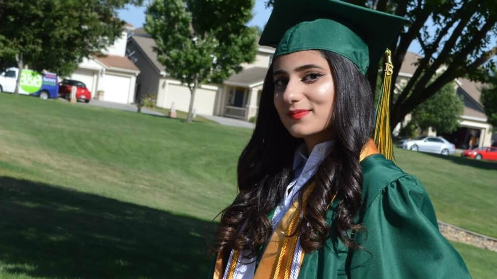 Woman in green graduation robe and cap smiles for camera, on residential street
