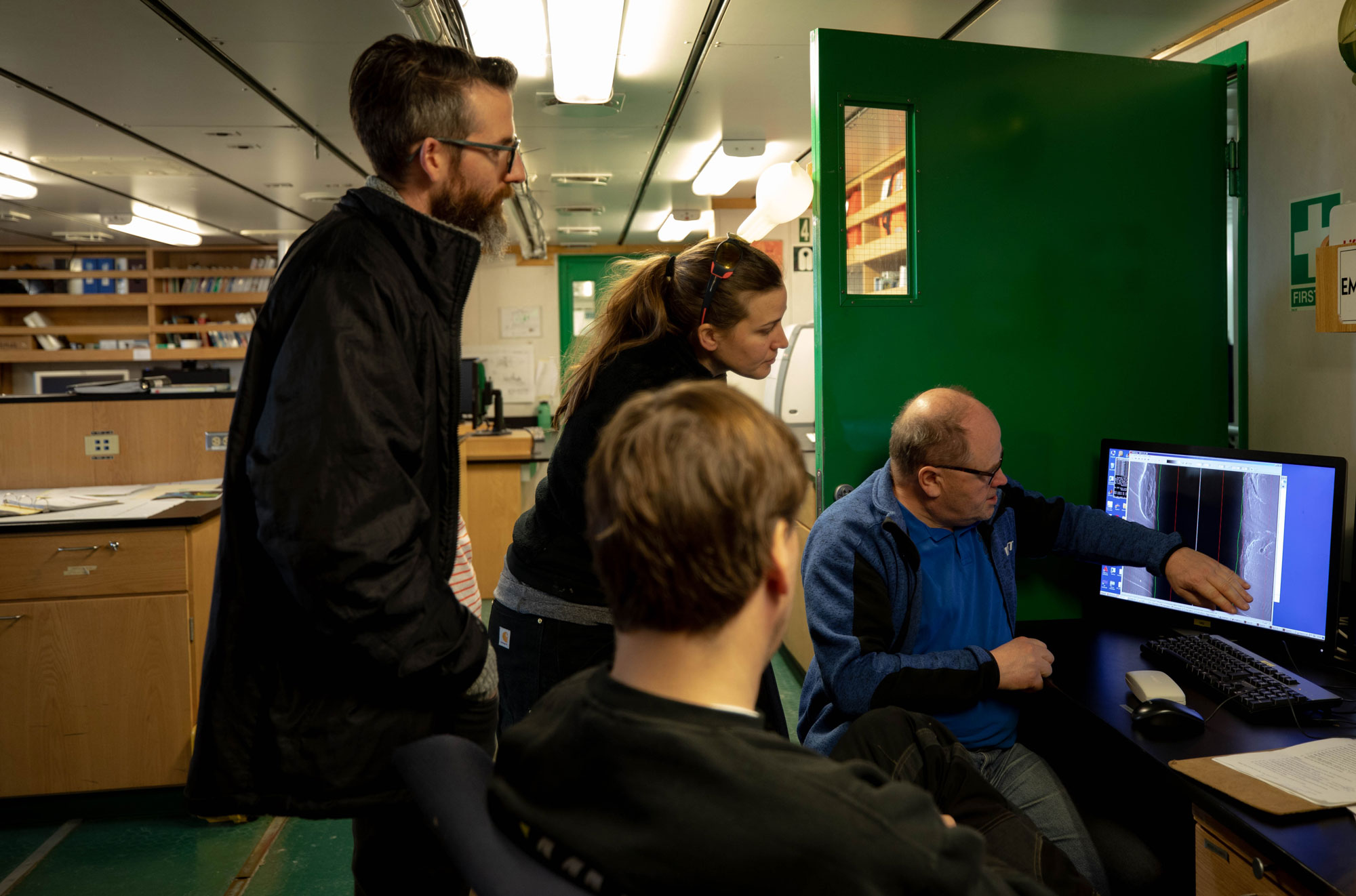 Scientists gathered in the ship’s lab to look at early images the Hugin captured of the seafloor near Thwaites. Scientists are shown tightly gathered around a computer.