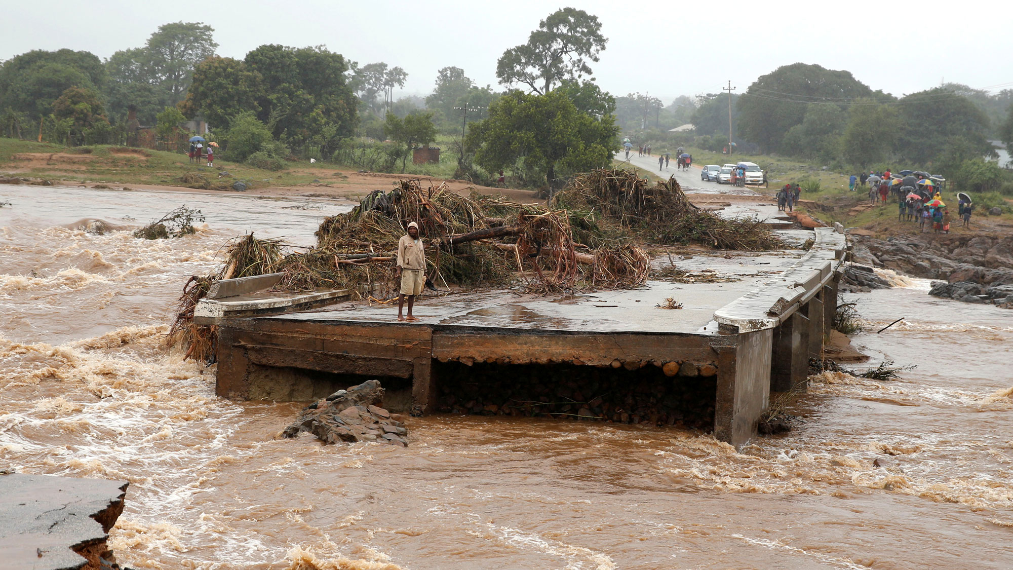 A man looks at a washed away bridge along Umvumvu river in Chimanimani, Zimbabwe. A man is shown standing with a hooded jacket looks at a washed away bridge.