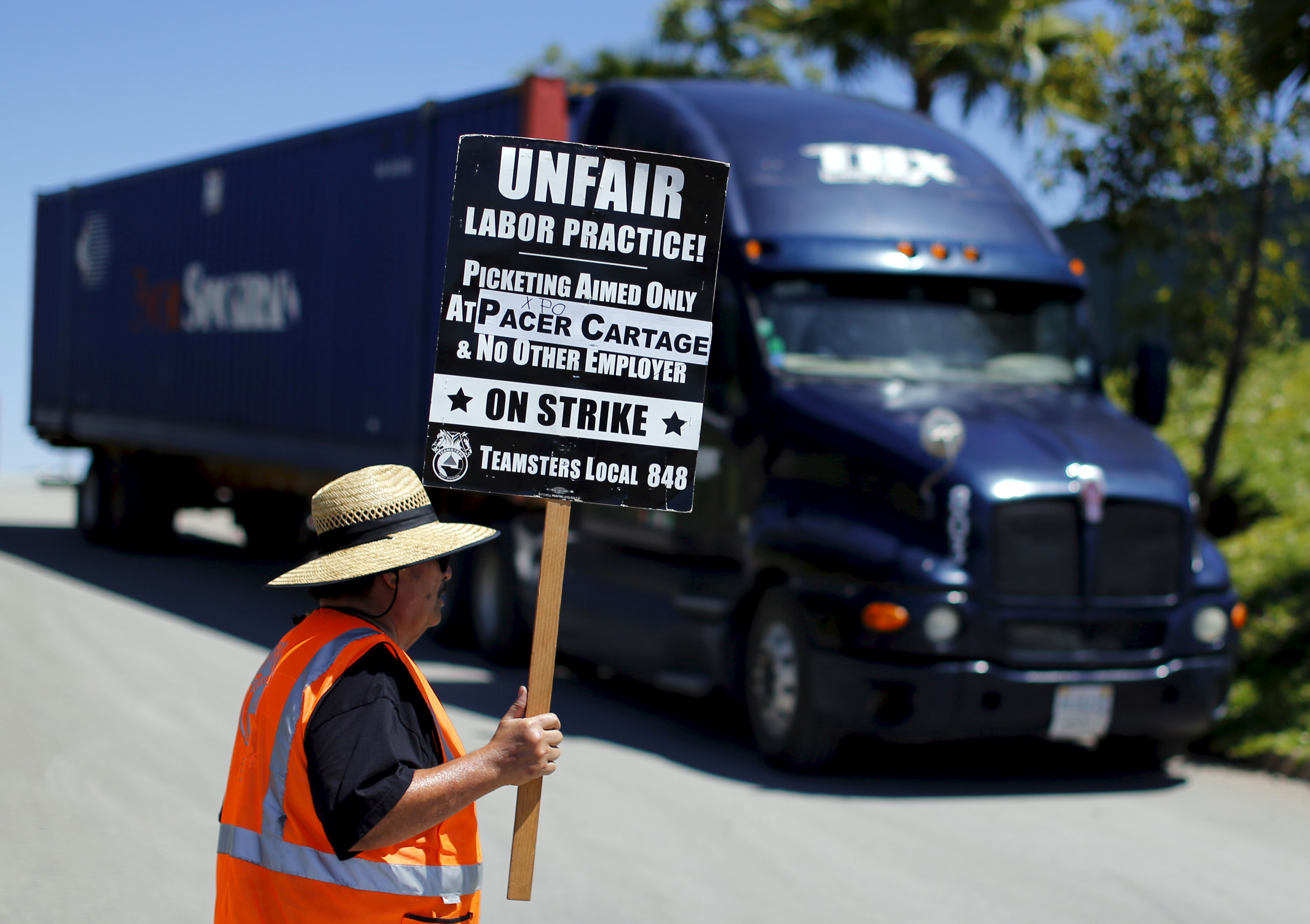 A striking truck driver walks a picket line at a Pacer Cartage facility in Otay Mesa, California. Drivers accused trucking companies of wage theft. A man next to a truck stands in an orange vest with a sign that says "unfair labor practice."