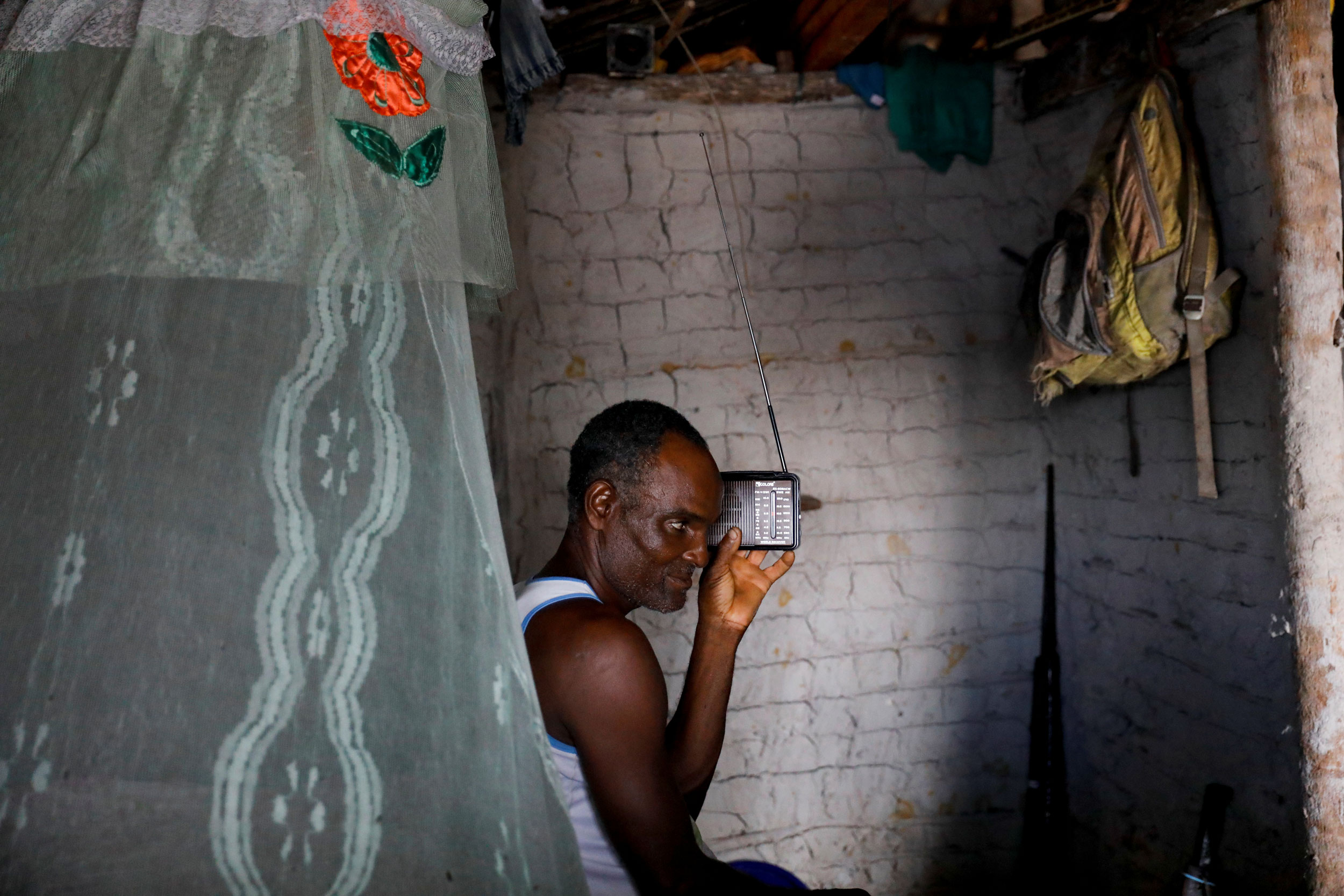 Cruz listens to the radio inside his house. A man is shown sitting with a small radio held next to his ear.