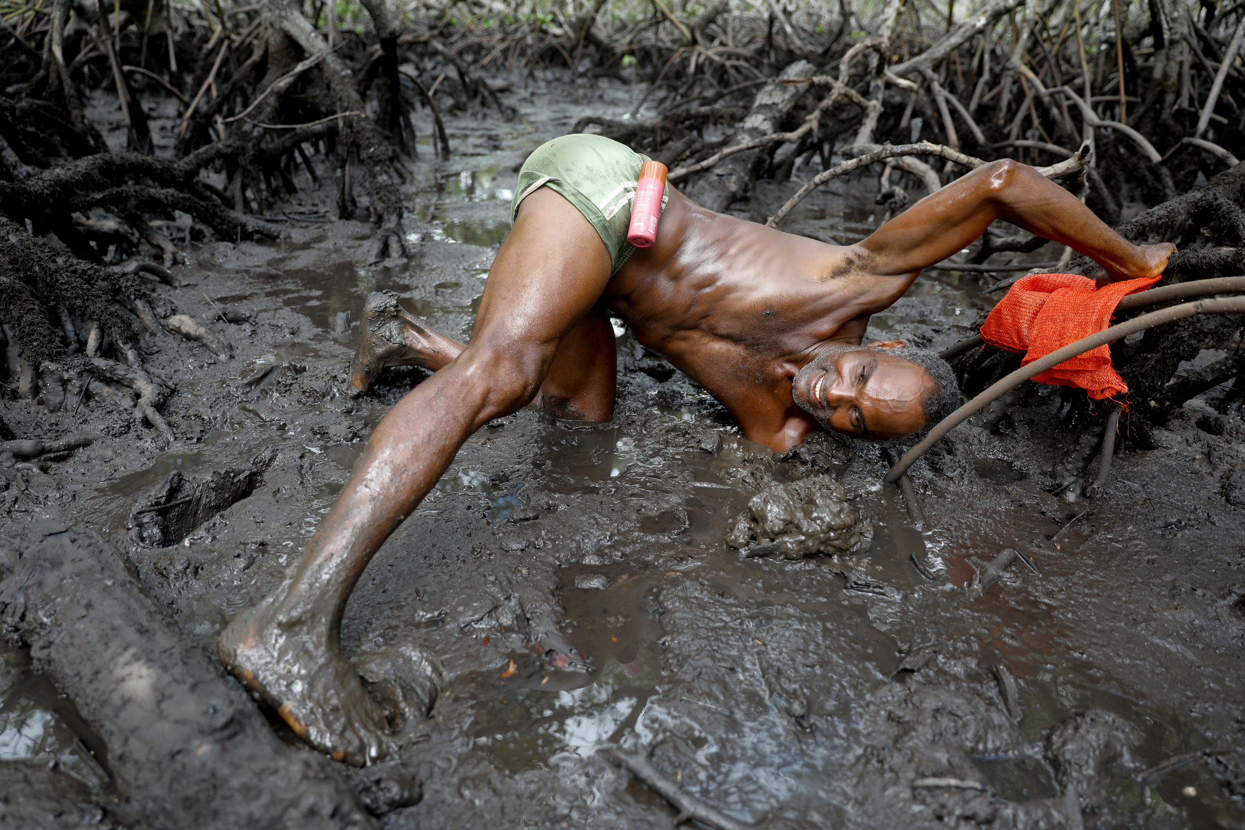 Cruz catches crabs. A man is shown laying on mud-covered mangroves wearing only green shorts with his left arm in the mud to his shoulder.