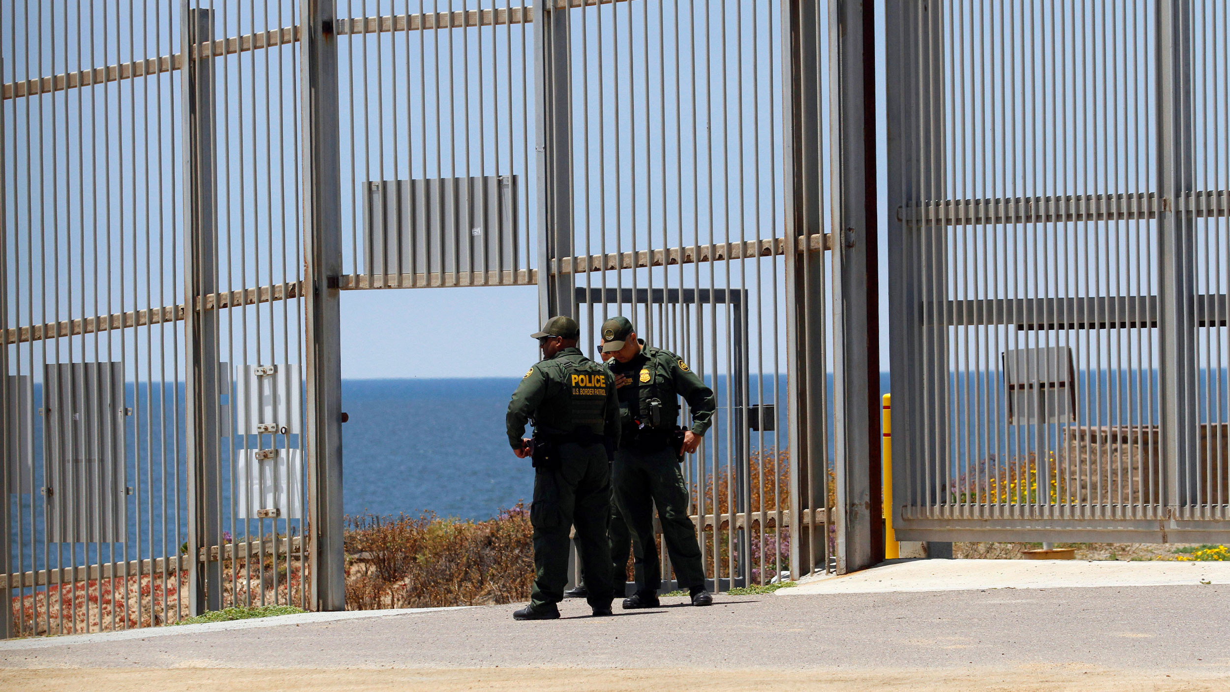 US Border Patrol agent are seen in the border fence between the United States and Mexico as photographed from Tijuana, Mexico, June 1, 2019. Two US Border Patrol agent are seen standing in uniform with their hands on their hips.