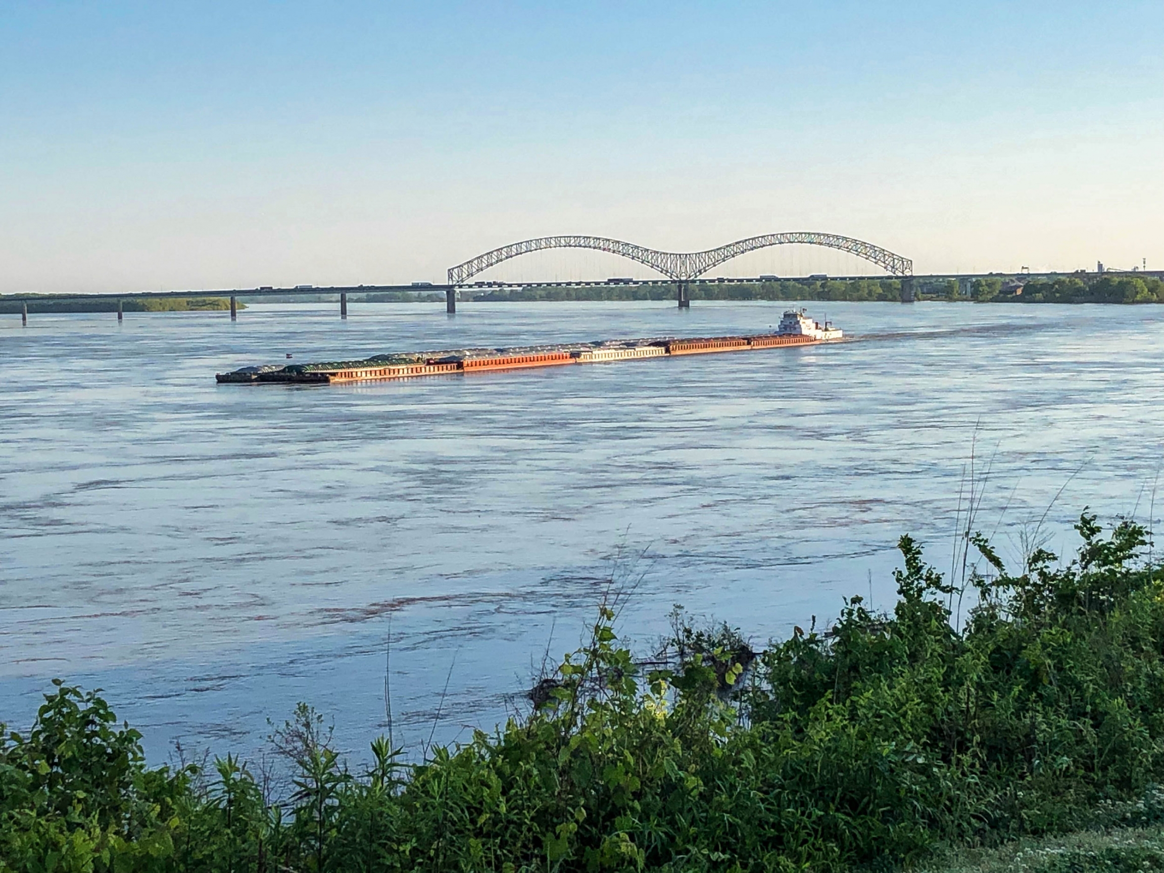 A barge carries goods down the Mississippi near Memphis, Tennessee. A barge is shown in the distance traveling on a blue river.