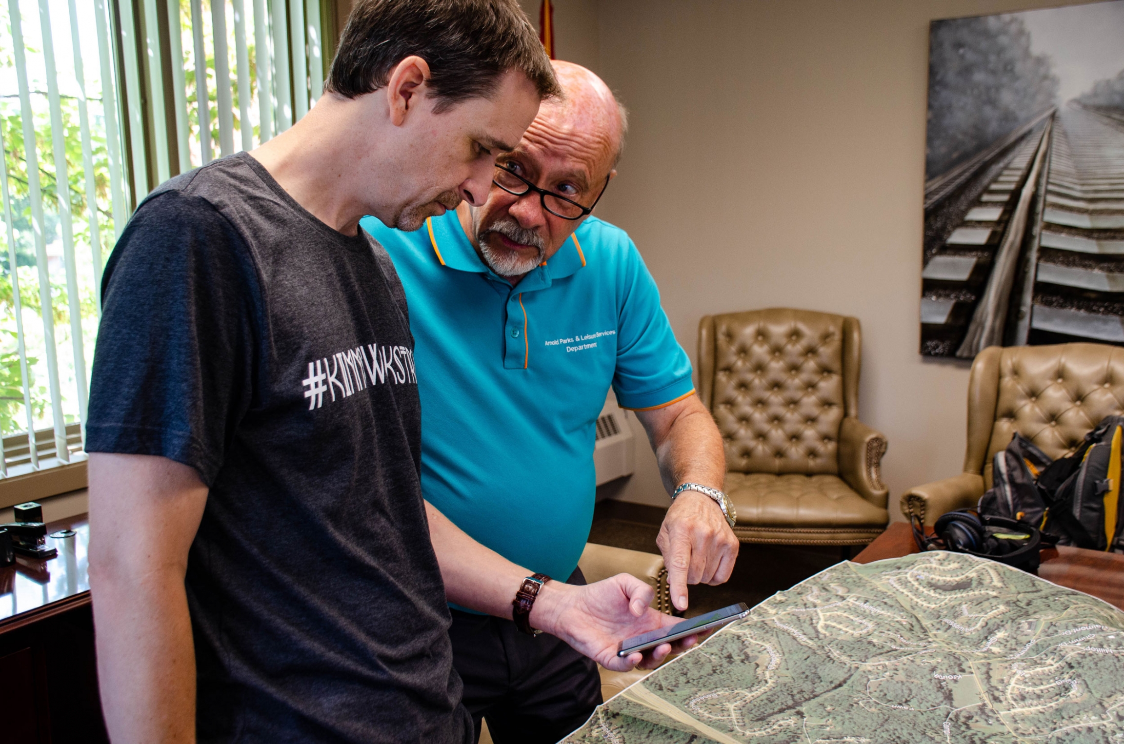 Colin Wellenkamp, executive director of the Mississippi River Cities and Towns Initiative, speaks with Ron Counts, mayor of Arnold, Missouri. Two men are shown speaking to each other with one holding a mobile phone.