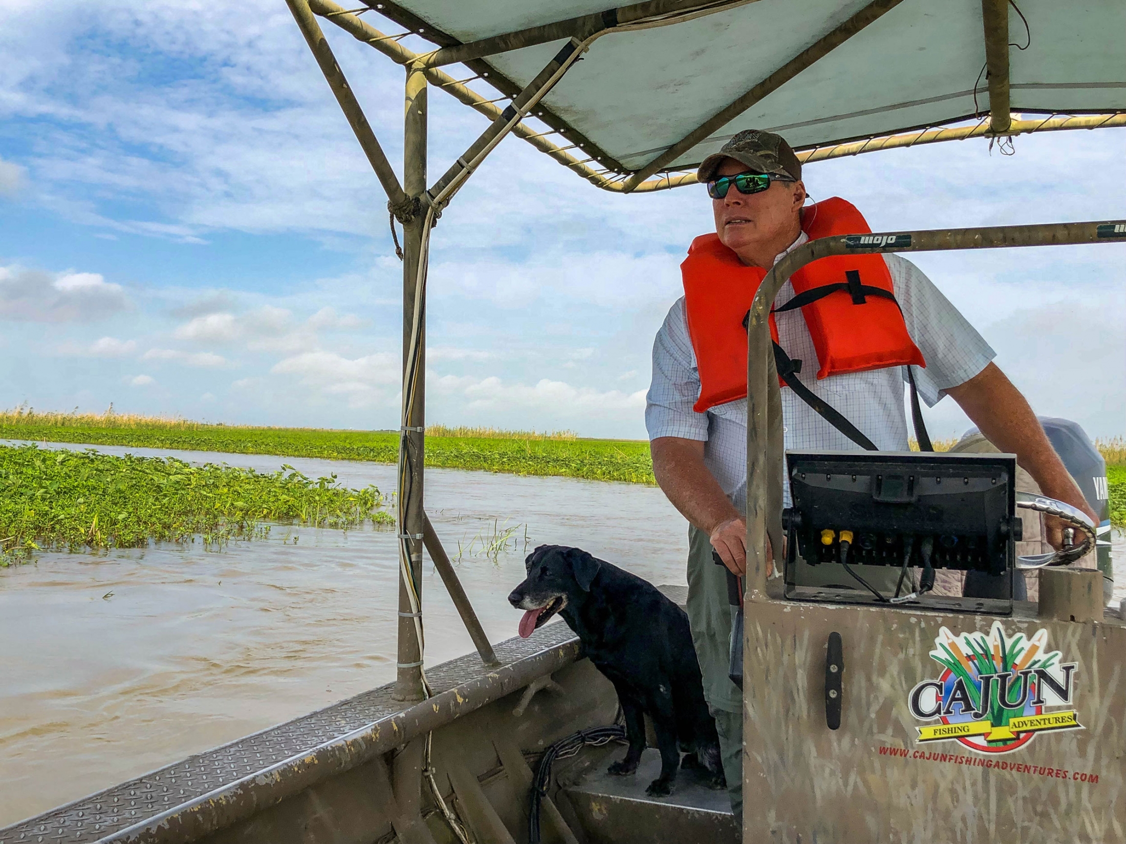 Ryan Lambert runs duck hunting and fishing tours, with his black lab Logan by his side, out of Buras, Louisiana, about 60 miles southeast of New Orleans. Ryan Lambert is shown piloting a flat bottom boat with a dark colored dog sitting next to him.