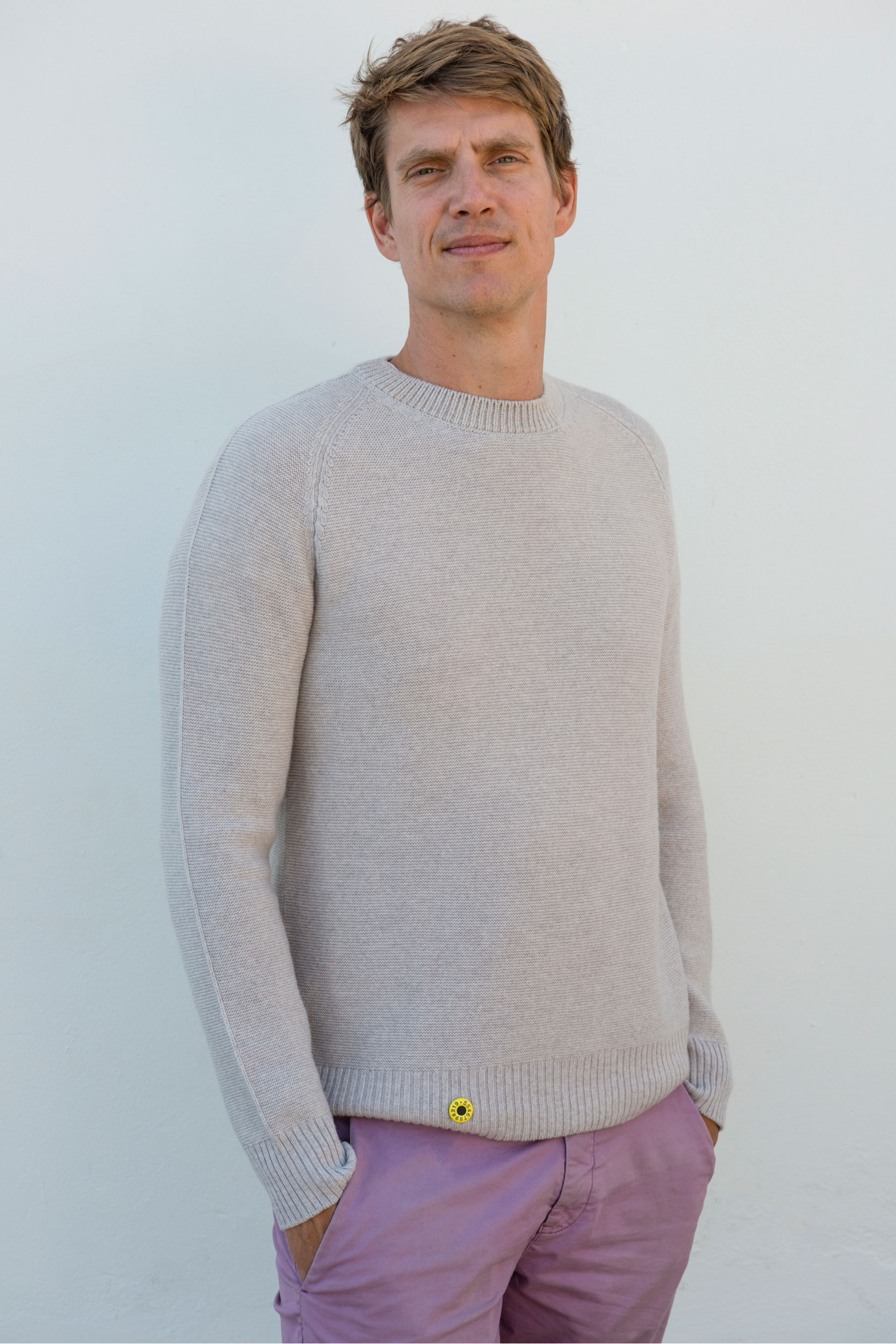 Sheep Inc. co-founder, Edzard van der Wyck, wears one of the brands sweaters. By scanning the small, yellow tag at the bottom, customers can learn the story of their garment. A white man in an off-white sweater and pink pants poses
