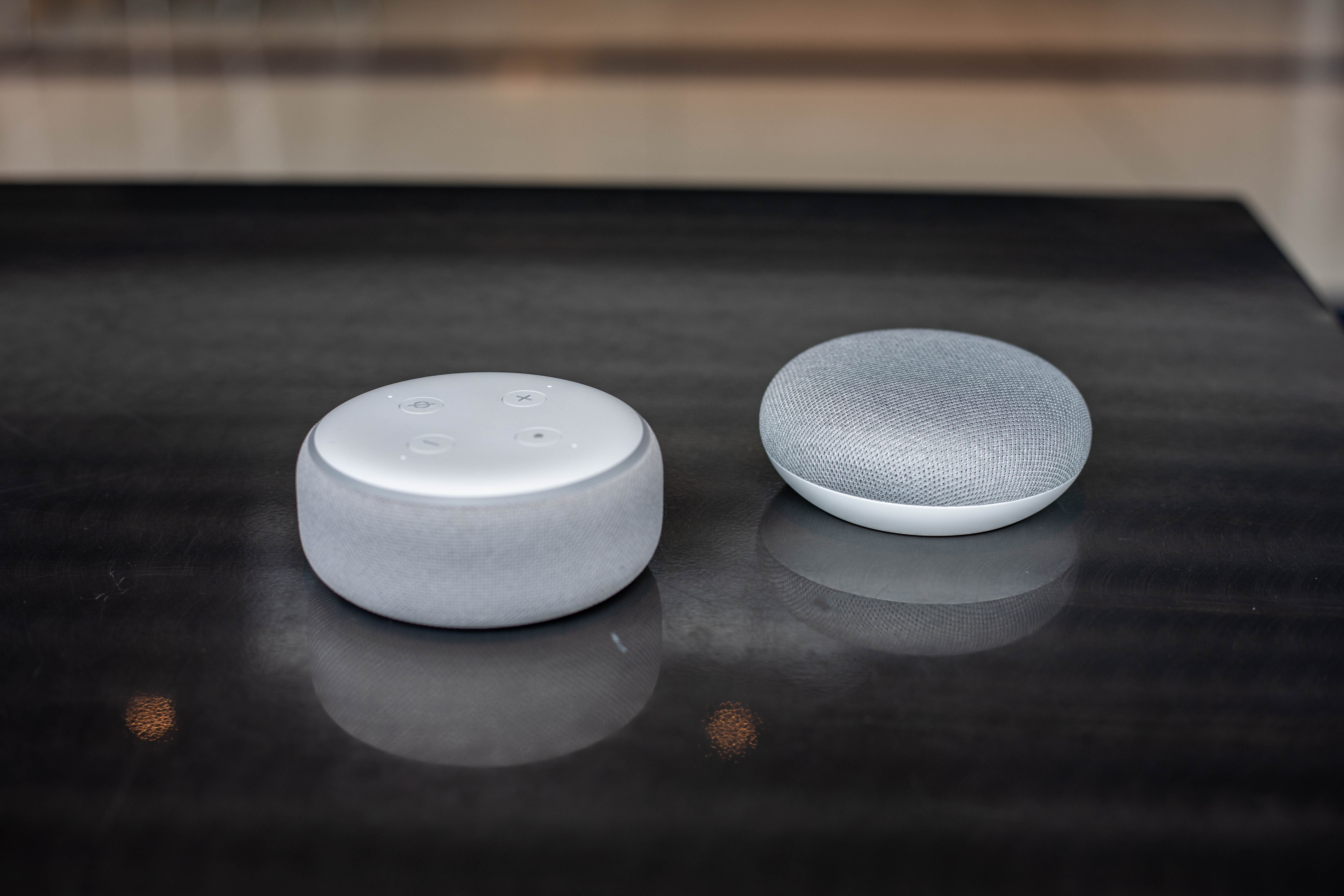 An Amazon Echo smart speaker sits next to a Google Home on a table.