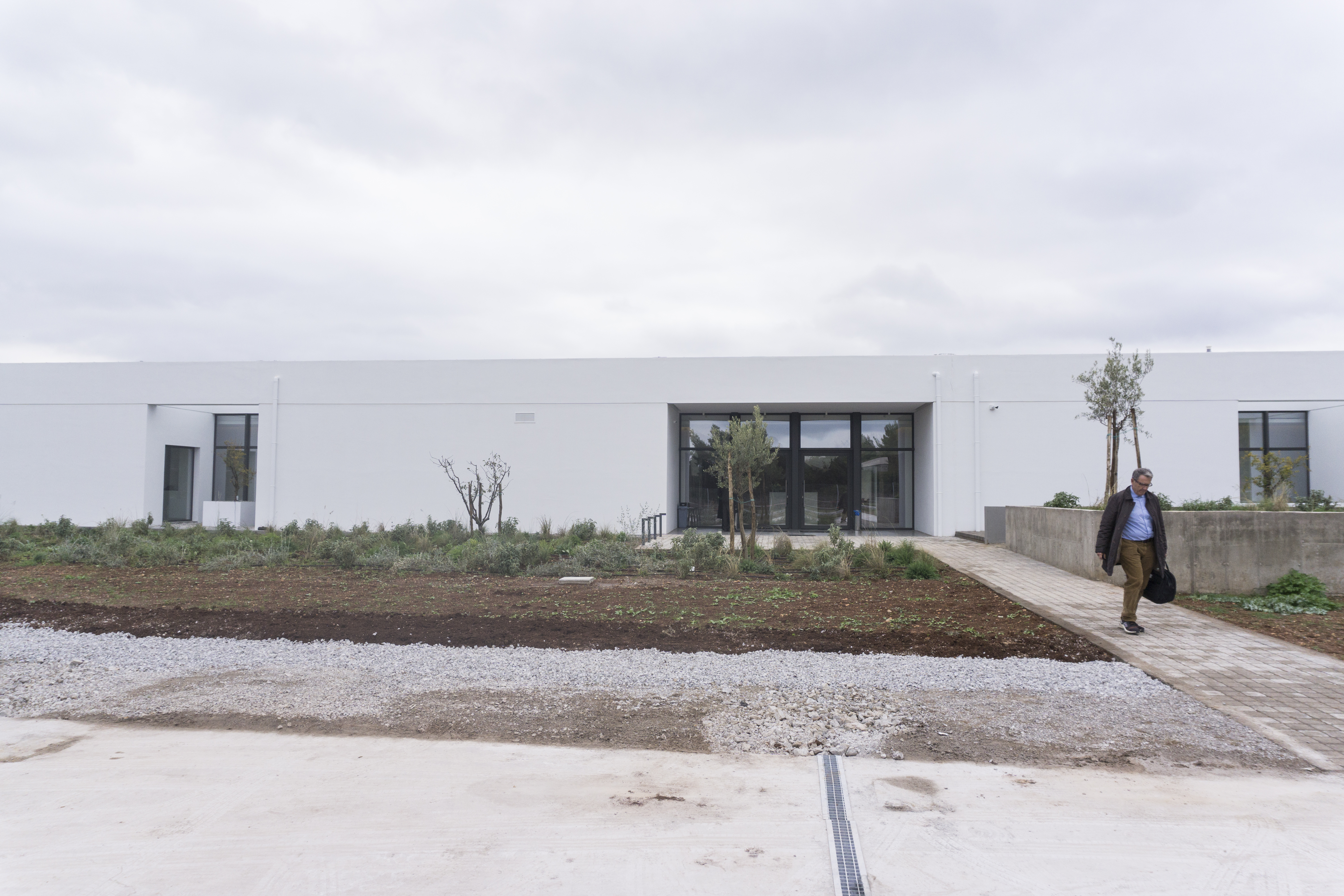 The Ritsona crematorium is located two hours away from central Athens, with a rose garden. A white building and sidewalk leading up to the doorway with a patch of green garden on the side.
