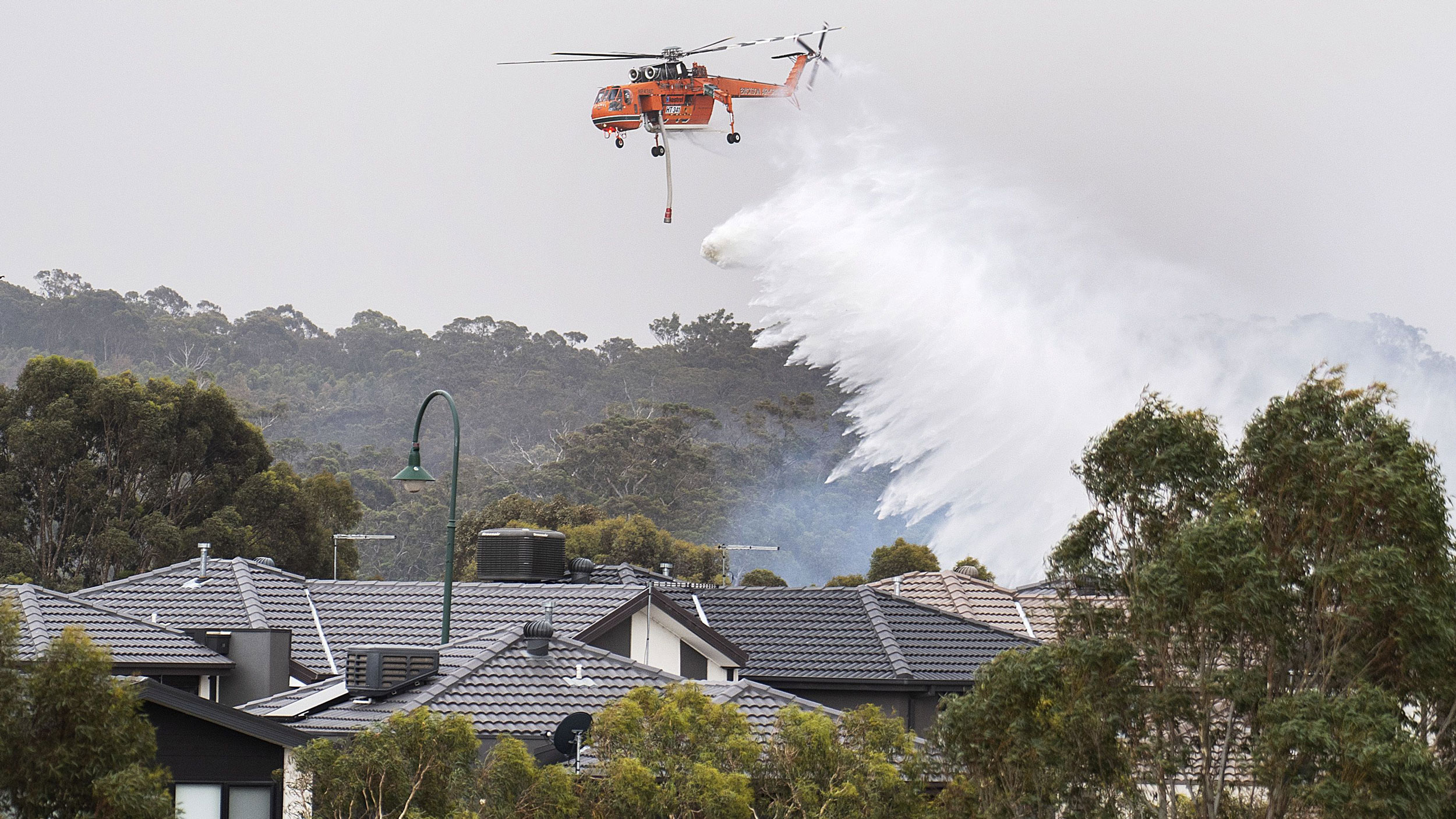 A helicopter drops water on a bushfire behind houses in Bundoora, Melbourne, Australia, Dec. 30, 2019. A large emergency helicopter is shown flying above several houses and dropping a large quantity of water.
