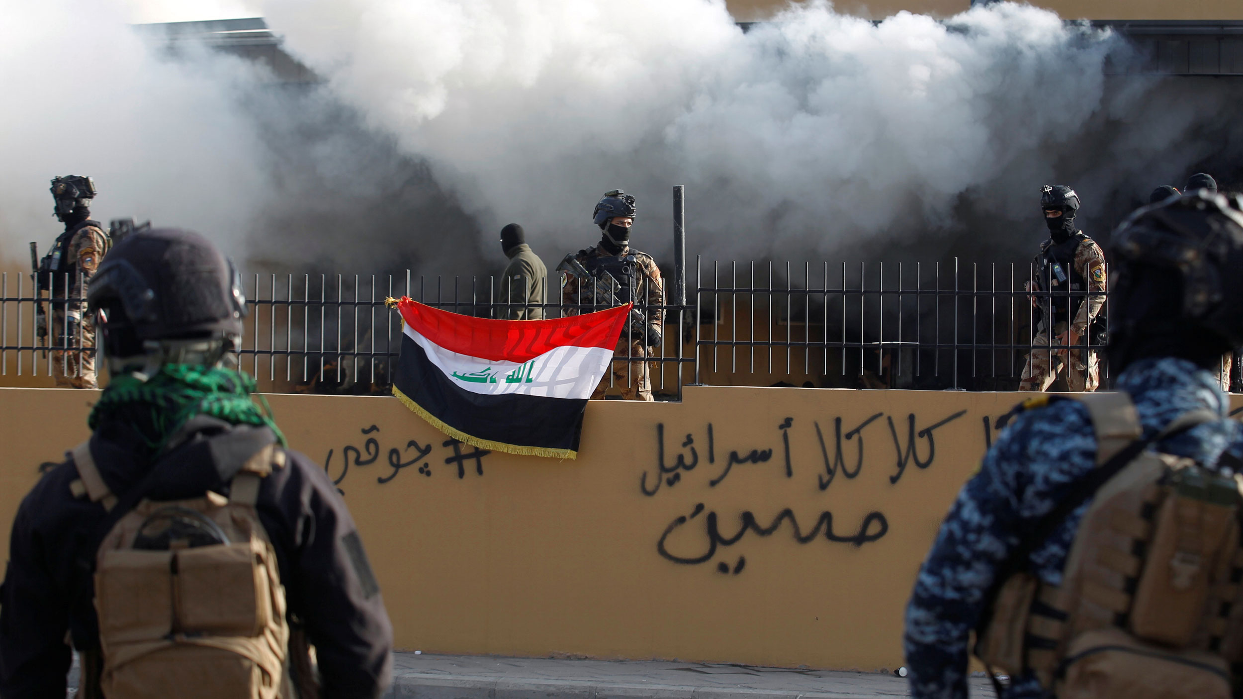 Members of Iraqi security forces are seen in front of US Embassy during a protest to condemn air strikes on bases belonging to Hashd al-Shaabi paramilitary forces, in Baghdad, Iraq, Jan. 1, 2020. Several Iraqi security authorities are shown standing guard behind a metal fence with smoke billowing out from the US Embassy behind them.