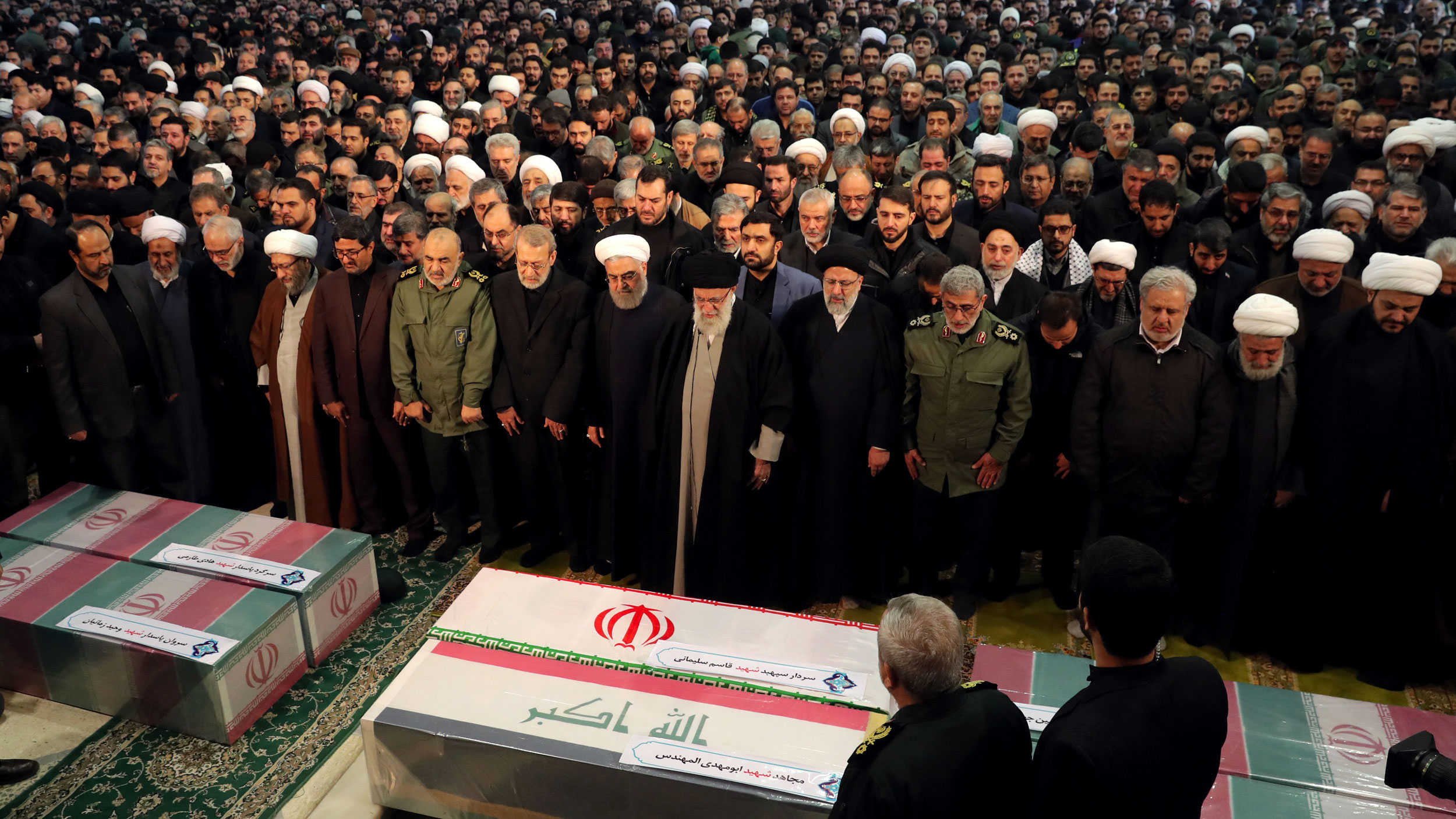 Iran's Supreme Leader Ayatollah Ali Khamenei and President Hassan Rouhani pray near the coffins of Iranian Maj. Gen. Qasem Soleimani, head of the elite Quds Force, and Iraqi militia commander Abu Mahdi al-Muhandis. Iran's Supreme Leader Ayatollah Ali Khamenei and President Hassan Rouhani are shown in front of a large group of people with their heads bowed and praying.