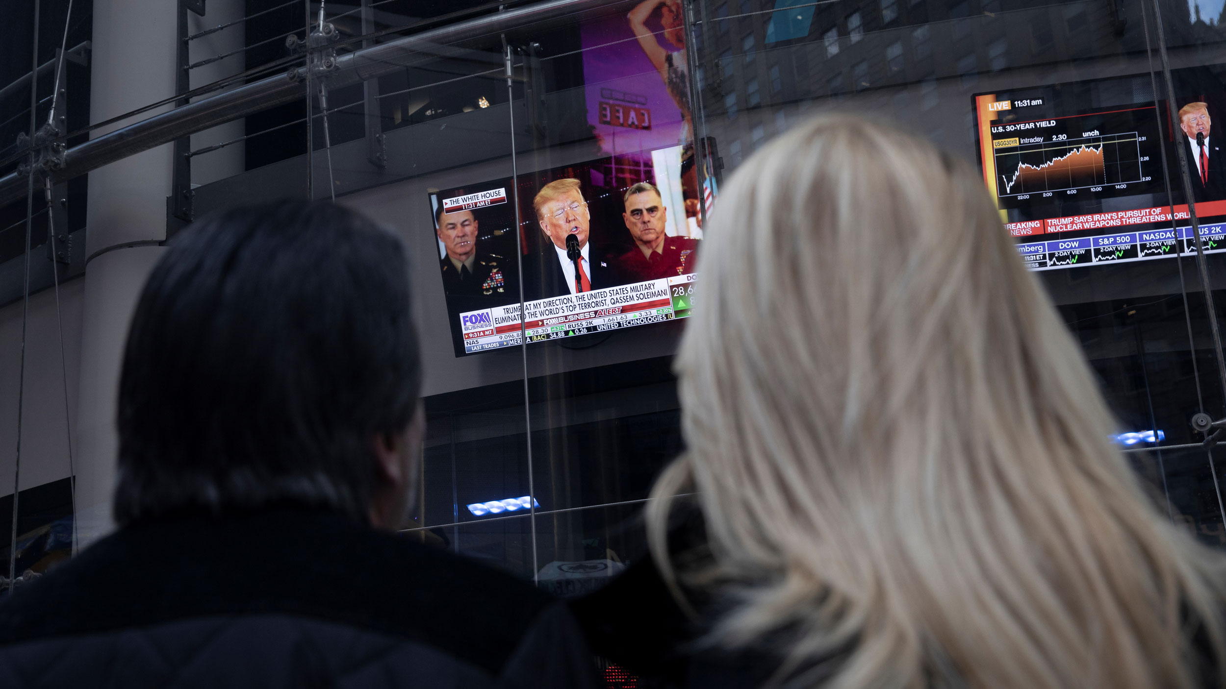 People watch as US President Donald Trump is seen on screens delivering a statement from the White House after Iran launched missile attacks on US-led forces in Iraq, in Times Square in New York on Jan. 8, 2020. Two people are shown from behind looking up at television monitors showing US President Donald Trump.