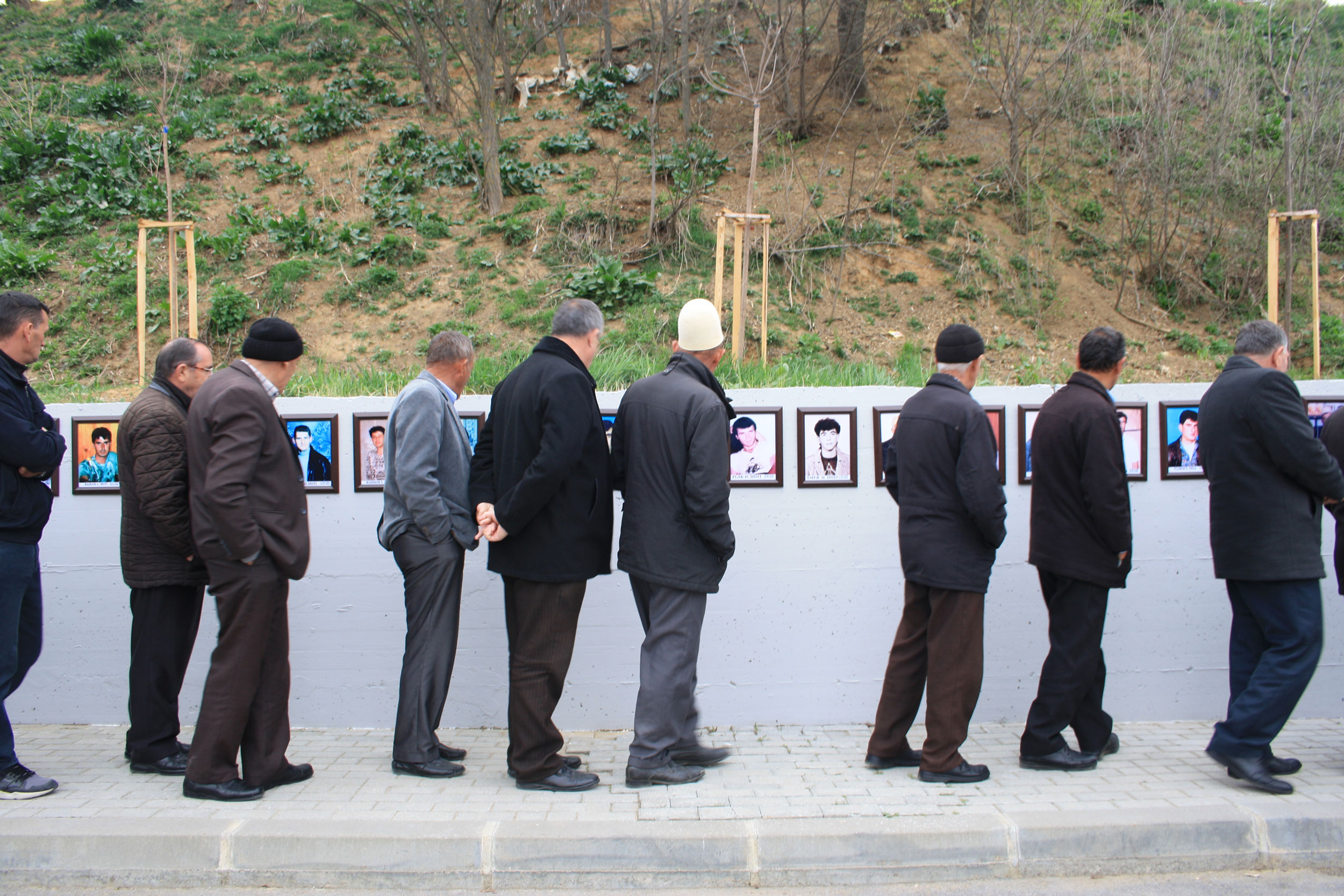 People attend a memorial on the anniversary of the massacre Velika Krusa, Kosovo. A line of men are shown standing next to a wall with photographs posted on it.