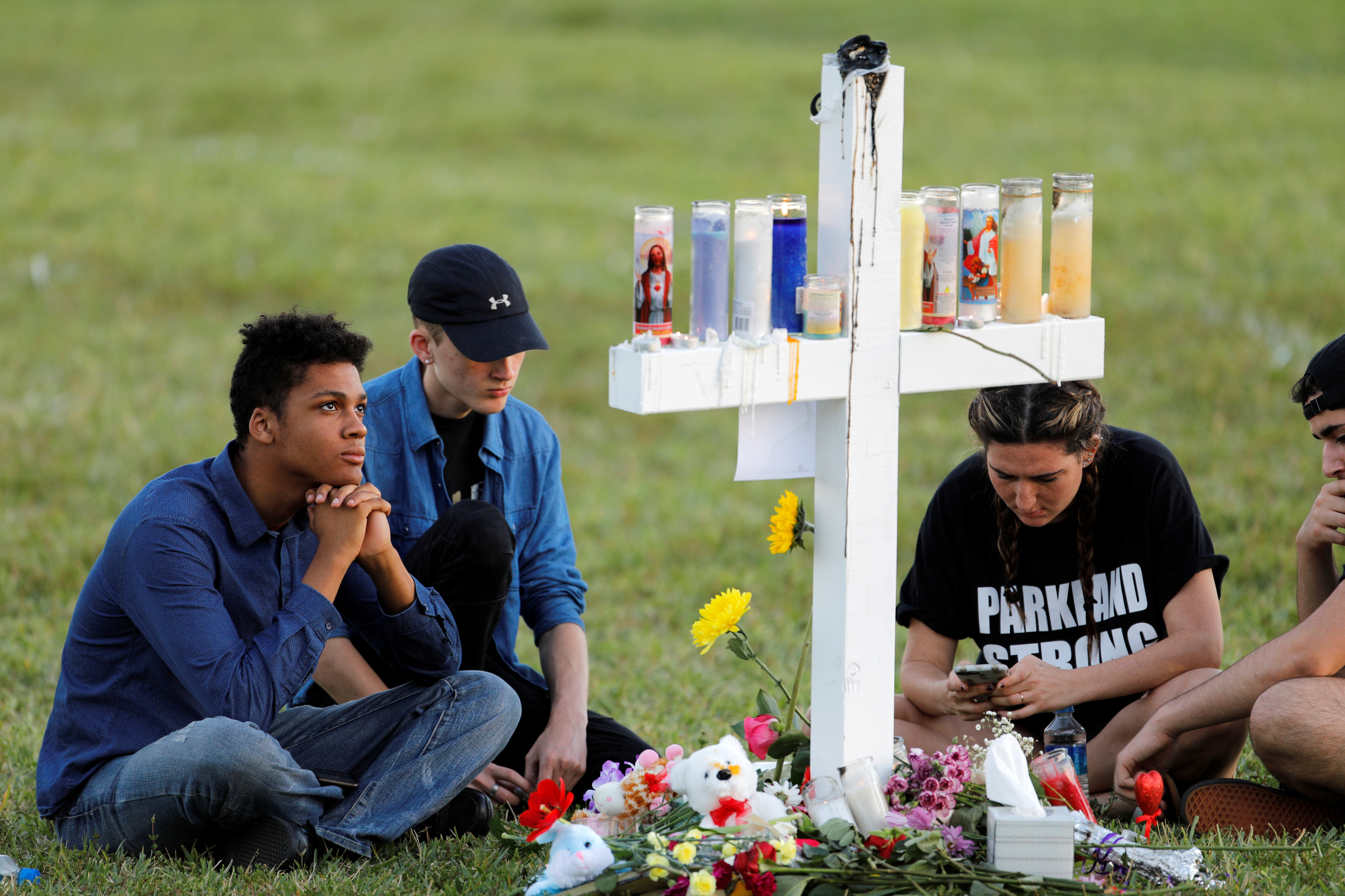 Mourners sit around one of 17 crosses at a memorial for the victims of the Parkland, FL, shooting. young people seated in grass around a cross