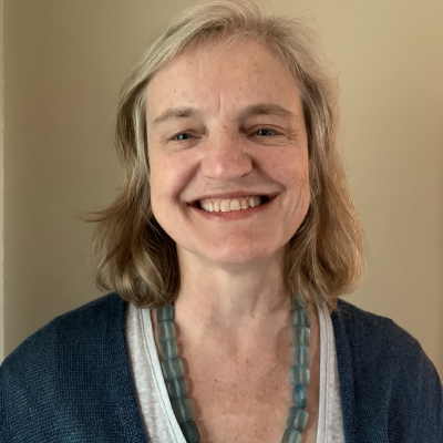 Carol Hills, The World's producer and host. a woman wearing a blue sweater and necklace smiles.