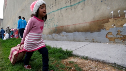 A Venezuelan migrant girl wearing a pink sweater with a heart on it, is shown heading to the exit of a makeshift camp with her belongings in Bogota, Colombia.