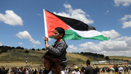 A young child sits on the shoulders of an adult waving a Palestinian flag under a blue sky