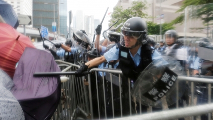 A police officer is shown with a helmet and clear face mask down, striking a person with an umbrella with his batton.