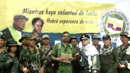 A group of men and women, some holding arms stand around a man reading from a document.