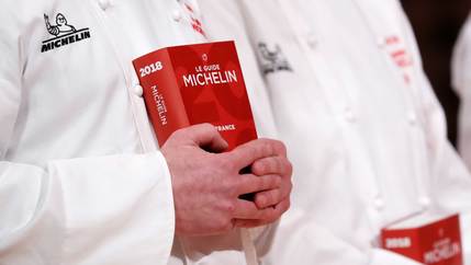 A pair of hands belonging to a person wearing a white chef coat holds a red 2018 Michelin Guide.