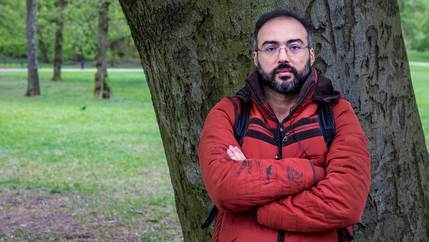 A man poses for a photo against a tree, wearing a red jacket