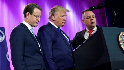 Three men in suits stand with their heads bowed.