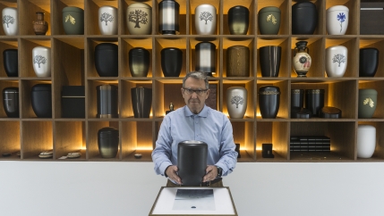 A man stands at a counter holding an urn with many urn designs behind him
