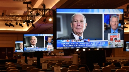An image of the face of Michael Bloomberg is shown on a large video monitor in an auditorium with empty seats.