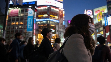 A large group of people are shown crossing an intersection, most wearing face masks, with brightly lit buildings in the background.