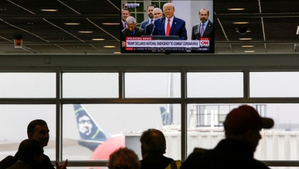 A television is shown hanging from the ceiling with US President Donald Trump displayed while several people are in shadow below.