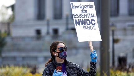 A woman in a mask and gloves holds a sign reading 