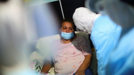 A woman is show sitting and wearing a medical safety mask and surrounded by health care workers wearing all blue protective gear.