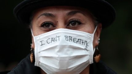 A close up photograph of a woman wearing a white protective face mask with the words, 