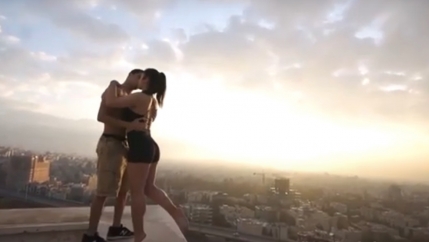 A man kisses a woman wearing blach shorts on a rooftop overlooking a pink skyline 