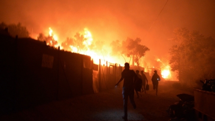 Several people are show running from a large fire engulfing several structures behind them.