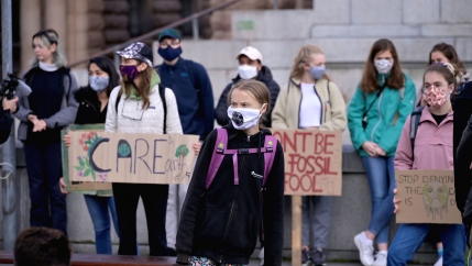 Swedish climate change activist Greta Thunberg is shown standing among a group of activists and wearing a purple backpack and a face mask.