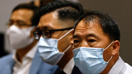 Three Asian men with short, dark hair wear face masks, the man in the middle wears glasses.  