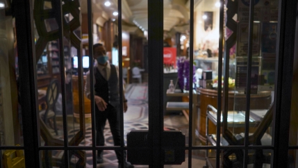 A man is shown wearing a face mask and tailored vest behind the closed-gated doors of a store front.