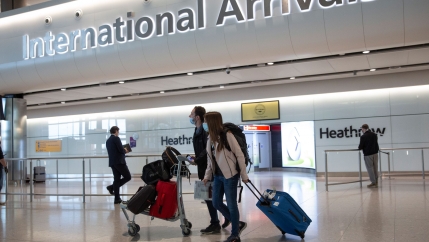 A pair of travelers are shown walking in the International Arrivals section of London's Heathrow Airport with one person pushing a cart carrying several bags.