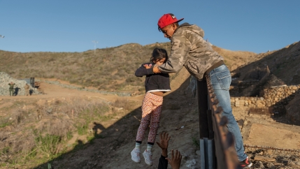 A child is passed over a border wall to the arms of a man wearing jeans, a red cap and tan sweatshirt. 