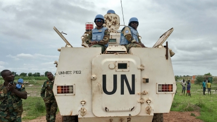 Two uniformed soldiers stand on top of a UN tank with two soldiers looking on and children in the background