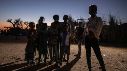 Several young people are shown in shadow standing with the sun setting behind them in the distance.