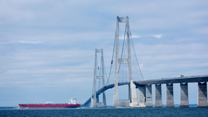 A large red oil tanker is shown in the distance passing underneath two tall bridge columns.